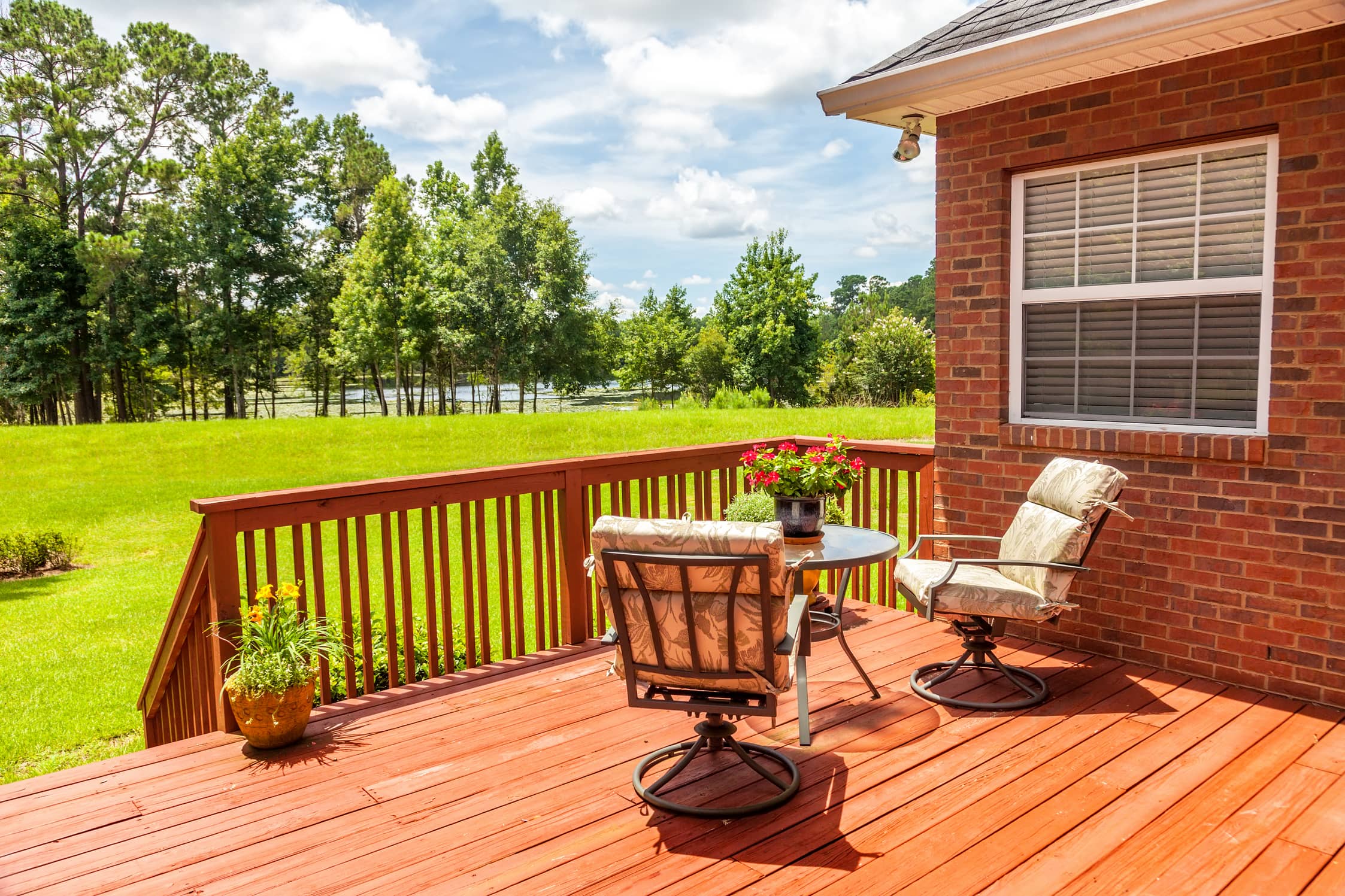 Image of a deck attached to a brick house with patio furniture next to a lawn and pond.